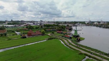 Yeşil tarlalarla çevrili nehir kenarındaki Hollanda yel değirmenleri ve bulutlu bir yaz gökyüzünün altında yürüyen turistler. Yeşil tarlalar ve huzurlu su manzaralı bir kırsal alan oluşturuyor..