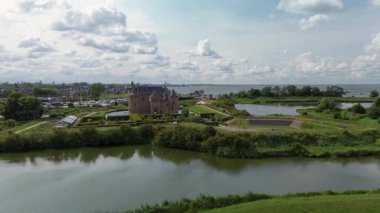 Hendek ve yeşil tarlalarla çevrili bir ortaçağ şatosunun havadan görünüşü, yürüyüş yolları, çadırlı bir yapı ve dramatik bulutlu bir gökyüzü altında yakınlardaki bir marina..