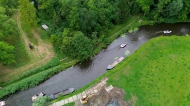 Kıyılar boyunca giden teknelerin, yemyeşil ağaçların, çimenli tarlaların ve kıyı şeridi yakınında küçük bir kazıcının bulunduğu dar bir nehrin havadan görünüşü. Huzurlu bir nehirde giden küçük bir tekne.