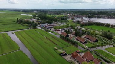 Yeşil tarlaları, dolambaçlı kanalları ve geleneksel evleri olan bir Hollanda kırsalının manzarası. İnsanlar yol boyunca yürürken nehir ve köy uzak ufka doğru uzanıyor..