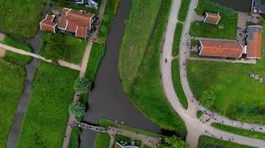 Nehir kenarındaki bir köyün hava manzarasında kırmızı çatılı evler, dolambaçlı yollar ve yeşil tarlalar görülüyor. Huzurlu bir günde insanlar kanal bankaları ve yollar boyunca yürüyor..