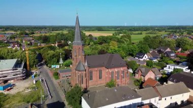 Evler, bahçeler ve yeşil ağaçlarla çevrili uzun bir çan kulesi olan tarihi bir kilisenin insansız hava aracı görüntüsü. Eski ve modern mimari ile harmanlanmış yollar ve bir inşaat sahası yakınlarda ortaya çıkıyor.