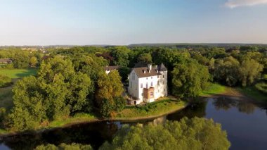 Sakin bir nehrin yakınında bulunan yemyeşil ormanlarla çevrili tarihi beyaz bir kale. Hava manzarası, temiz bir gökyüzünün altında ağaçlar, tarlalar ve uzak köylerle huzurlu bir kırsal bölgeyi vurguluyor..