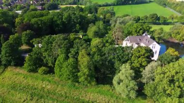 Sakin bir nehrin yakınında bulunan yemyeşil ormanlarla çevrili tarihi beyaz bir kale. Hava manzarası, temiz bir gökyüzünün altında ağaçlar, tarlalar ve uzak köylerle huzurlu bir kırsal bölgeyi vurguluyor..