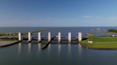 Beton sütunlu bir deniz barajının havadan görünüşü, deniz ile iç sular arasında akan suyu kontrol etmek, ufukta yollar, yeşil alanlar ve yelkenli teknelerle çevrili..