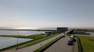 Deniz savunması olan kıyı yolu ufka doğru uzanır, sakin sular ve yeşil karayla sınırlıdır, kıyıya yakın arabaları ve uzak denizde yelkenlileri vardır..