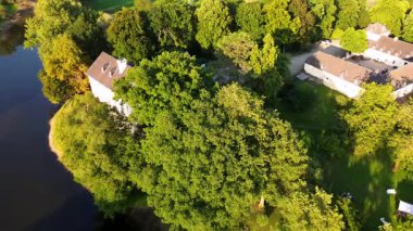 Sakin bir nehrin yakınında bulunan yemyeşil ormanlarla çevrili tarihi beyaz bir kale. Hava manzarası, temiz bir gökyüzünün altında ağaçlar, tarlalar ve uzak köylerle huzurlu bir kırsal bölgeyi vurguluyor..