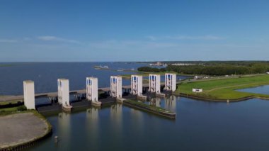 Beton sütunlu bir deniz barajının havadan görünüşü, deniz ile iç sular arasında akan suyu kontrol etmek, ufukta yollar, yeşil alanlar ve yelkenli teknelerle çevrili..