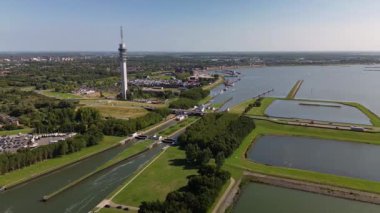 Yüksek bir iletişim kulesi nehir kenti üzerinde yükselmekte, kanallar, köprüler, botlar ve yeşil manzaralarla modern altyapıyı doğal çevreyle harmanlamaktadır..