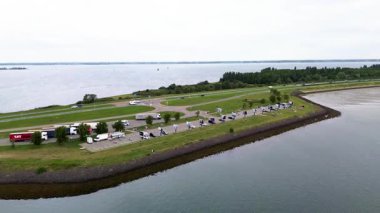 Deniz kenarındaki otoban dinlenme alanının hava aracı görüntüsü, sahil şeridi boyunca park halindeki kamyonlar, arabalar ve kamyonetler, yeşillik, karayolu ağı ve sakin sularla çevrili..