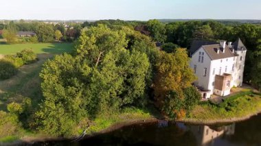 Sakin bir nehrin yakınında bulunan yemyeşil ormanlarla çevrili tarihi beyaz bir kale. Hava manzarası, temiz bir gökyüzünün altında ağaçlar, tarlalar ve uzak köylerle huzurlu bir kırsal bölgeyi vurguluyor..