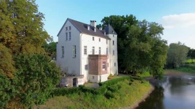 Sakin bir nehrin yakınında bulunan yemyeşil ormanlarla çevrili tarihi beyaz bir kale. Hava manzarası, temiz bir gökyüzünün altında ağaçlar, tarlalar ve uzak köylerle huzurlu bir kırsal bölgeyi vurguluyor..