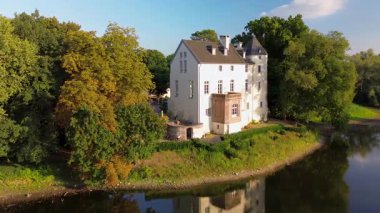 Sakin bir nehrin yakınında bulunan yemyeşil ormanlarla çevrili tarihi beyaz bir kale. Hava manzarası, temiz bir gökyüzünün altında ağaçlar, tarlalar ve uzak köylerle huzurlu bir kırsal bölgeyi vurguluyor..