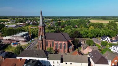 Amsterdam, Kuzey Hollanda, Hollanda - 09.06.2025: Küçük bir Avrupa kasabasındaki evler, yeşillikler ve tarlalarla çevrili sivri uçlu yüksek Gotik tarzı bir kilisenin havadan görünüşü.