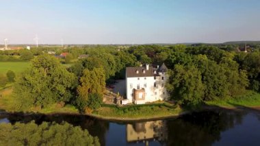 Sakin bir nehrin yakınında bulunan yemyeşil ormanlarla çevrili tarihi beyaz bir kale. Hava manzarası, temiz bir gökyüzünün altında ağaçlar, tarlalar ve uzak köylerle huzurlu bir kırsal bölgeyi vurguluyor..