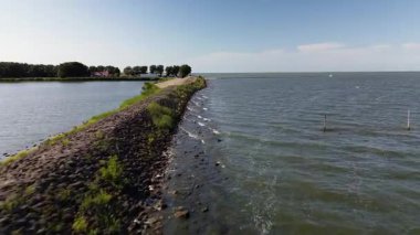 Su kütleleri arasında uzanan bir barajın manzaralı görüntüsü, bir tarafta dalgalar, diğer tarafta sakin su, yeşillik ve birkaç ev ile çevrili..
