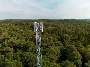 Uzun bir telekom kulesi ve toprak erişim yolu olan sık ormanın havadan görünüşü teknoloji ve doğal manzaranın kesişimini vurguluyor..