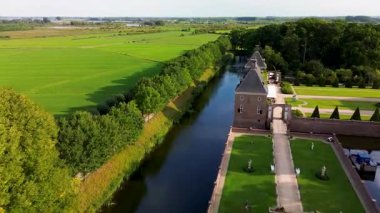 Hendek, orman ve tarlalarla çevrili taş bir kalenin havadan görünüşü. Bir köprü girişe götürür, manikürlü bahçeler ve sakin kırsal alanlar.