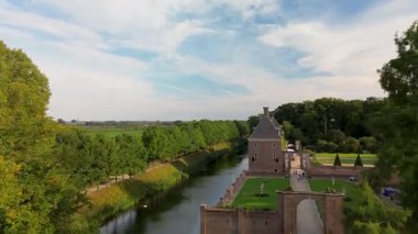 Hendek, orman ve tarlalarla çevrili taş bir kalenin havadan görünüşü. Bir köprü girişe götürür, manikürlü bahçeler ve sakin kırsal alanlar.