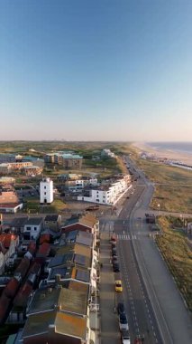 Beyaz bir kilisesi, kırmızı çatılı evleri ve manzaralı bir avlusu olan kıyı kasabasının havadan görünüşü. Kumsal, kum tepeleri ve sahil kulübeleri kıyı boyunca uzanır. Mimariyi doğayla harmanlarlar..
