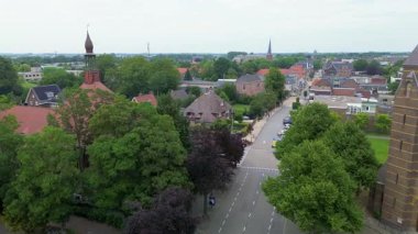Kırmızı kiremitli ve sazdan çatılı evleri olan banliyö caddesi, park edilmiş arabalar, bahçeler ve uzak kilise kulesi mimari çeşitlilik ve düzenli planlama sergiliyor..