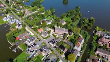 Göl kenarındaki yüksek kilise, rıhtım, tekne ve ağaç kaplı göl köyünün hava manzarasında sakin mimari, su girişi ve yeşil topluluk planlaması sergileniyor..