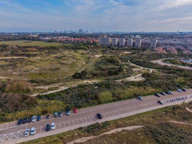 Yeşil parkın dolambaçlı yolları ve yol kenarındaki park alanının havadan görünüşü, doğayla metropol arasındaki kontrastı vurgulayan yoğun kentsel ufuk çizgisine geçiş.