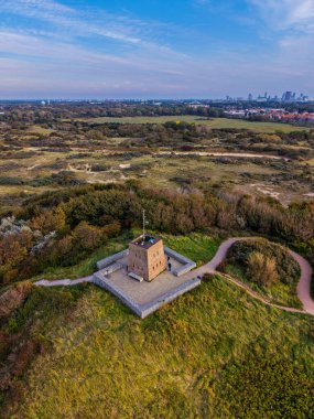 Antenli izole tuğla binanın hava manzarası, çimenli arazide taş duvarla çevrili doğal açıklık ile uzak kentsel ufuk çizgisi ve dolambaçlı yollar..