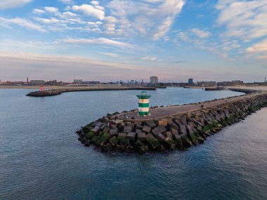 Yeşil çizgili deniz feneri, açılı beton bloklar ve deniz altyapısını manzaralı gökyüzü altında kentsel zemin ile harmanlayan uzak şehir manzaralı kıyı dalgakıranı..