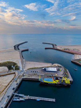 Kum, dalgakıran, kumlu plajlar ve gün doğumu veya günbatımı gökyüzü altında kıyı altyapısı ile çevrili, RESEARCH tesisi yakınındaki sarı mavi araştırma gemisinin havadan görüntüsü..