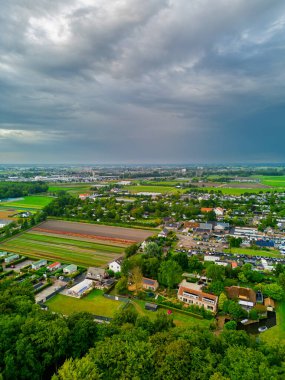 Ağaçlarla kaplı evler, ekin tarlaları ve uzak kasaba katmanlı arazi kullanımının dramatik bulutlu gökyüzü altında hava manzarası.