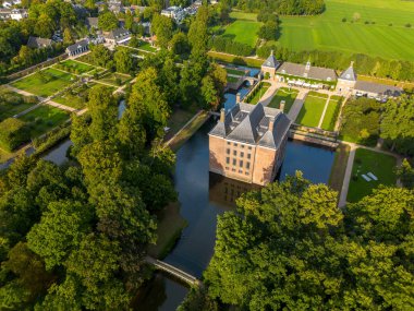Bahçeleri, köprüsü ve çevresi ağaçlar, su hatları ve sıcak güneş ışığı altındaki yakınlardaki bir köyle çevrili tarihi kale manzarası..