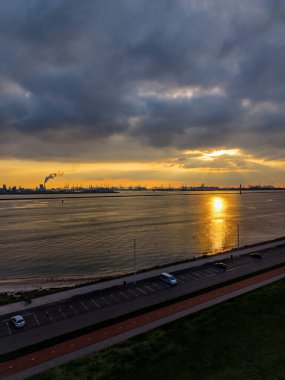 Kıyı günbatımının havadan görünen altın ışığı, park halindeki arabaların ve yaya yolunun yakınındaki sakin suya yansıyor. Ufukta vinçlerden ve duman bacalarından oluşan endüstriyel ufuk çizgisi var..