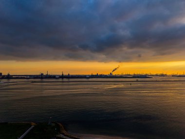Gün batımında kıyı sanayi bölgesinin havadan görünüşü sakin su, kumlu plaj ve vinçler, bacalar ve çarpıcı bir çevresel tabloda çarpıcı turuncu gökyüzü ile ön plan kontrastı..