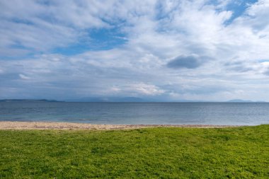 Green grass at empty sandy beach. Winter day, heavy fluffy cloudy sky background meets rippled dark blue sea. Greek seascape. Space.