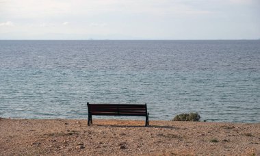 Lonely empty old wooden bench on soil over the beach. Winter sunny day, calm blue sea, cloudy sky background. Greek seascape. 