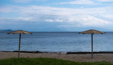 Two wooden parasol on an empty sandy beach. Winter day, blue rippled sea and cloudy sky background. Seascape from Greece.