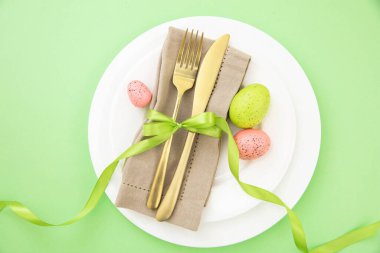 Paskalya sofrası, Paskalya yumurtası süslemesi, peçete ve altın çatal bıçak takımı beyaz tabaklarda, pastel yeşil arka plan
