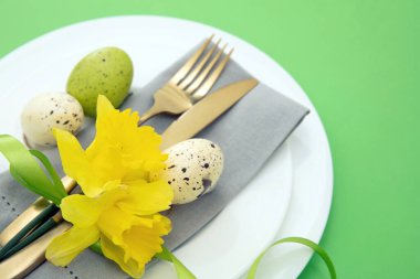 Paskalya sofrası, bahar çiçekleri ve yumurtalar süsleme, beyaz tabaklarda peçete ve altın çatal bıçak takımı, pastel yeşil arka plan