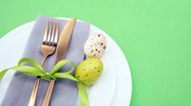 Paskalya sofrası, Paskalya yumurtası süslemesi, peçete ve altın çatal bıçak takımı beyaz tabaklarda, pastel yeşil arka plan