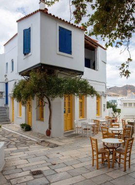 Yunanistan. Tinos Adası Cyclades. Pyrgos köyünde sarı pencereli geleneksel bir kafe. Asfaltlı bahçede boş sandalye ve masa