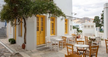 Yunanistan. Tinos Adası Cyclades. Pyrgos köyünde sarı pencereli geleneksel bir kafe. Asfaltlı bahçede boş sandalye ve masa