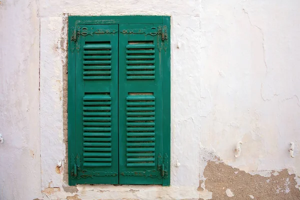 Old fashioned window with closed green wooden shutter on faded, peeled white wall background. House facade, Cyclades island, Greece. Space