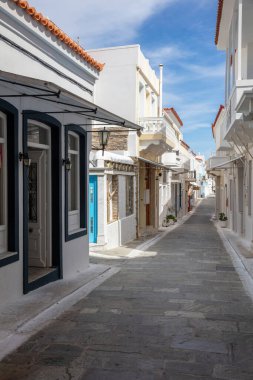 Yunanistan, Kiklad. Andros Adası, Chora Kasabası. Parke taşı boş dar bir sokak, beyaz boyalı ev ve dükkan duvarı, mavi gökyüzü arka planı. Dikey