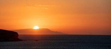 Yunan adası üzerinde gün batımı, Cyclades, Yunanistan. Altın güneş tepe siluetinin arkasında saklanıyor. Gökyüzü turuncu, sarı. Uçsuz bucaksız Ege Denizi. Pankart