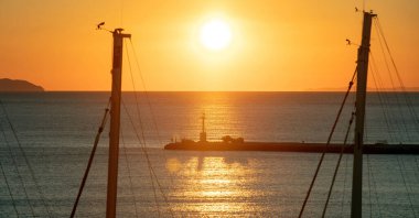 Günbatımı Tinos Adası, Cyclades, Yunanistan 'da. Gemi direği, altın güneş, gökyüzünü renklendiren ve Ege Denizi 'ni ışıldatan fener siluetinin görüntüsü.