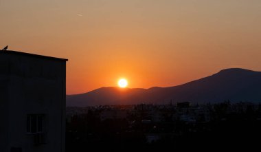 Şehrin üzerinde gün batımı. Altın güneş, turuncu ve kırmızı arkaplanlı gökyüzünü renklendirir. Alacakaranlığa hayran olan bir bina ve güvercin silueti..