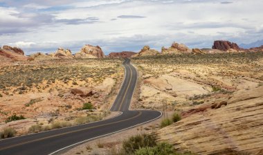 Fire State Park Vadisi Nevada, ABD. Boş kıvrımlı otoyol, Amerikan çölü, düşük bitki örtüsü, kırmızı kayalık oluşum, bulutlu gökyüzü arkaplanı.