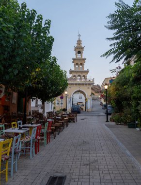 Evangelistra Kilisesi Palaiochora Chania, Girit Adası, Yunanistan. Asfalt caddedeki açık kafe, öğleden sonra kemerli çan kulesi girişi. Dikey