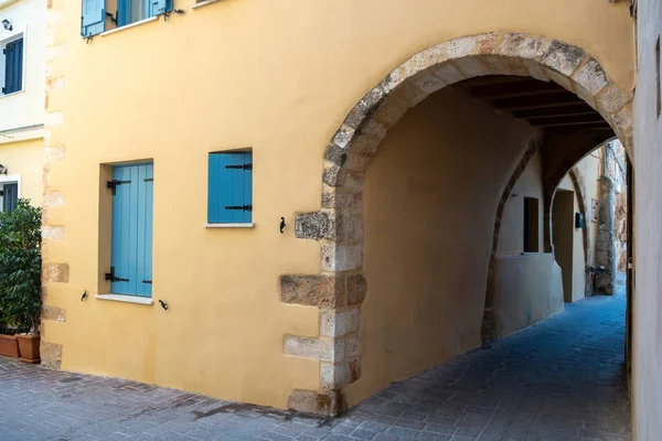 Girit adası, Chania Old Town, Yunanistan. Dar, boş asfalt sokağı kaplayan kemerli taş duvarın üzerindeki mavi panjurlu bina.. 